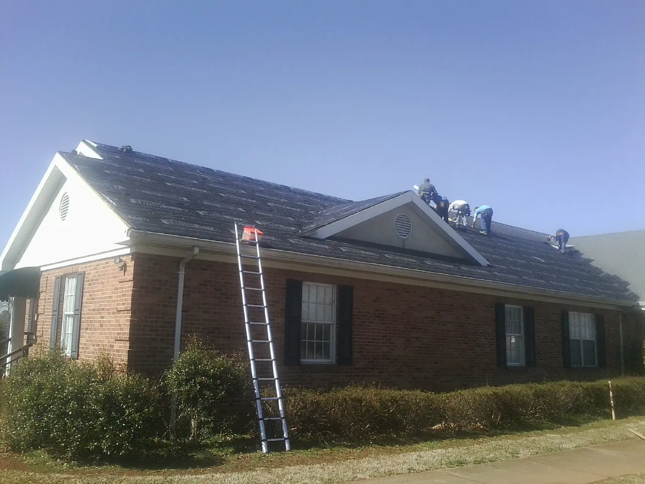 Crew tearing shingles off a brick ranch roof with a ladder set — Emergency Roof Repair work by our crew in Gracey