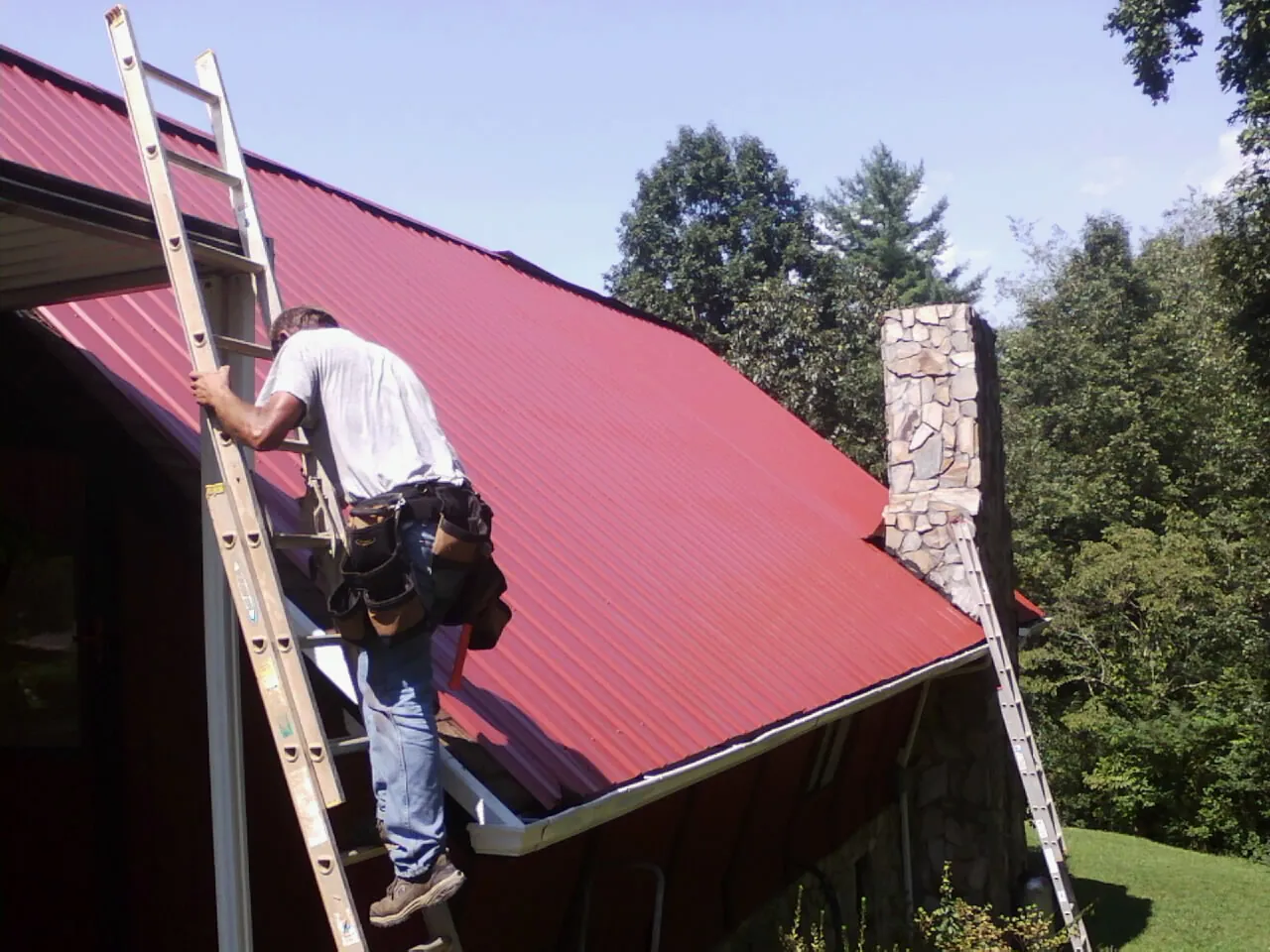 Roofer on a ladder beside a red metal roof and stone chimney — Soffit & Fascia Repair by Roofing Brothers in Gracey